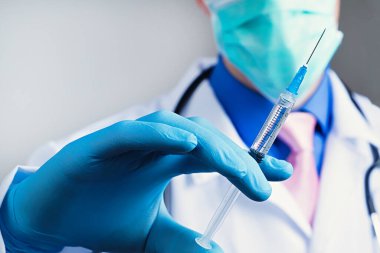 Plastik mavi eldivenli tıbbi şırıngaya yakından bak. El giydirilmiş, göğsü beyaz tıbbi bornozlu, steteskoplu, bulanık bir arka planı var.