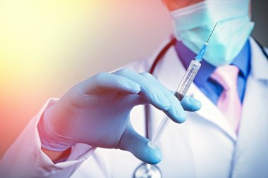 Plastik mavi eldivenli tıbbi şırıngaya yakından bak. El giydirilmiş, göğsü beyaz tıbbi bornozlu, steteskoplu, bulanık bir arka planı var.