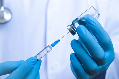 Şırınganın ve cam ampulün görüntüsünü kapat. Mavi lastik eldiven giymiş, beyaz bornoz giymiş, arka planda bulanık doktor.
