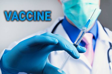 Plastik mavi eldivenli tıbbi şırınganın yakın görüntüsü, bulanık arka planda steteskoplu beyaz tıbbi bornozlu adamın göğsü. Aşı kavramı