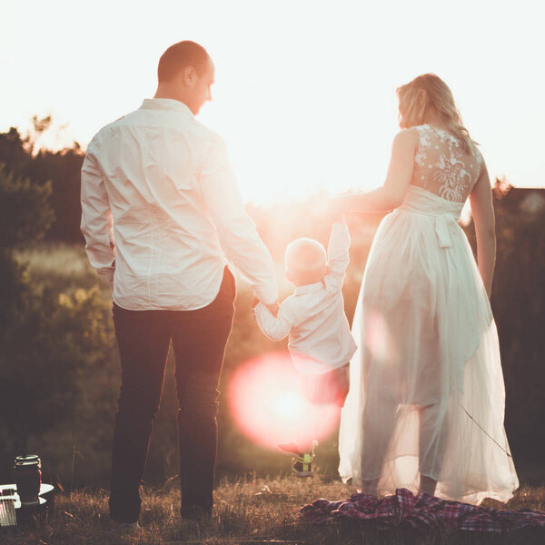 Little family romantic picnic scene outdoor in sunset. Parents holding hands of little toddler boy between them.