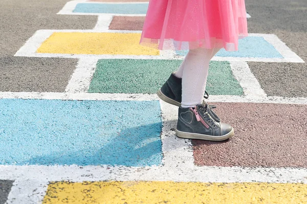Closeup of girl\'s legs in a pink dress and hopscotch drawn on asphalt. Child playing hopscotch on playground outdoors on a sunny day. outdoor activities for children.