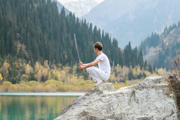 A wise man sits on a stone and holds a sword in his hands. Mountain Lake background