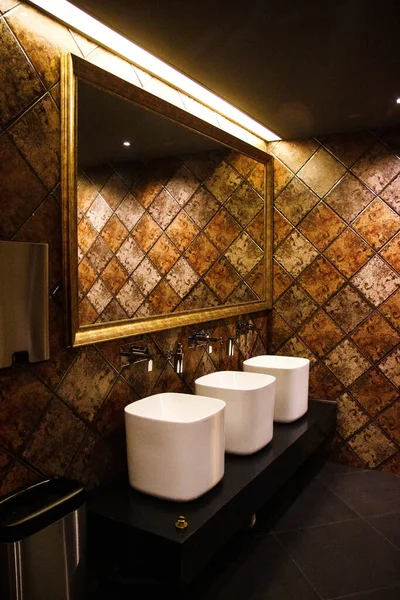 Golden brown design water closet room with bronze color tile on a wall and big mirror three sinks and paper tissue dispenser