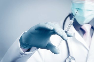 Gazlı bez maskeli bir doktor ve elinde mavi kauçuk eldiveninde şırınga tutan bir doktor. Sağlık ve aşı kavramı