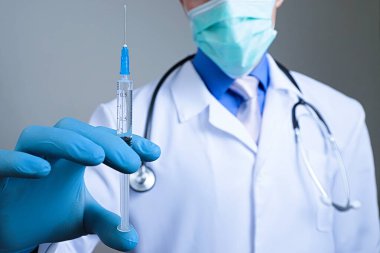 Plastik mavi eldivenli tıbbi şırıngaya yakından bak. El giydirilmiş, göğsü beyaz tıbbi bornozlu, steteskoplu, bulanık bir arka planı var.