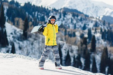Genç kadın snowboardcu kayak pistinde yürüyor omuzlarında kayak tahtası, kameraya bakıyor, boşluğu kopyalıyor.
