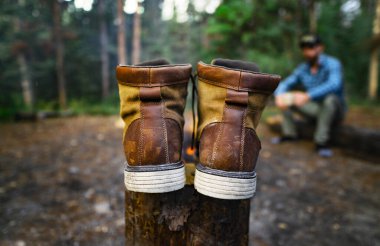 Deri süet botlar, arka planda bir adamla vahşi doğada ateşin yanında oturur.