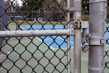 Tenis kortunun kapısı asma kilitli ve arka planda tenis ağı var..