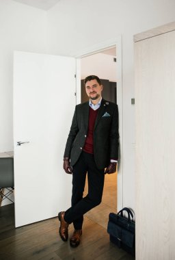 Şık giyimli, sakallı bir iş adamı şık bir takım elbise, kahverengi ayakkabılar, kravat, eldivenler, pahalı bir dairede poz veren bir adam.