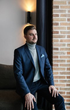 Şık giyimli, sakallı bir iş adamı şık bir takım elbise, kahverengi ayakkabılar, kravat, eldivenler, pahalı bir dairede poz veren bir adam.