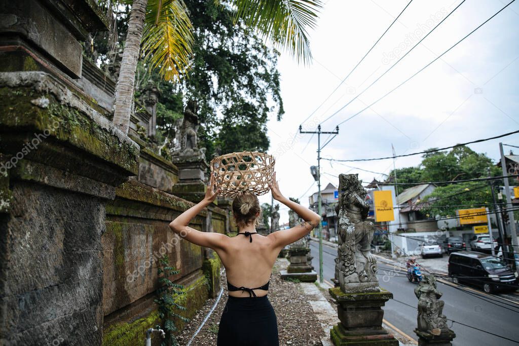 blonde blogger girl poses at an ancient temple in the jungle with a wicker basket while traveling in Indonesia on the island of Bali