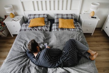 Çizgili cüppeli ve damalı pantolonlu şık bir adam yatak odasının iç kısmında sarı yastıklı gri çarşaflar üzerinde poz veriyor.