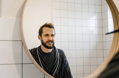 Sakallı, bornozlu şık bir adam banyoda dişlerini fırçalar ve aynaya bakar.