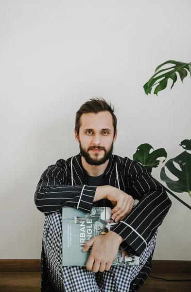 stylish man with a beard in a striped robe and checkered pants posing in the interior of his bedroom next to a green plant