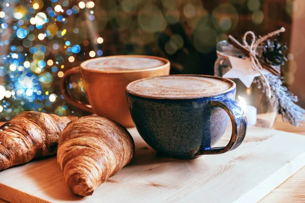 Perfect breakfast of croissants and cappuccino in a cafe. Blue and yellow ceramic cups of cappuccino and croissants on wooden table with Christmas lights and bokeh background. Mockup for your design.
