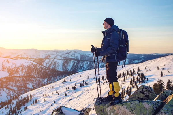 Mannen vandrare stående på snötäckta bergstopp i solnedgången — Stockfoto