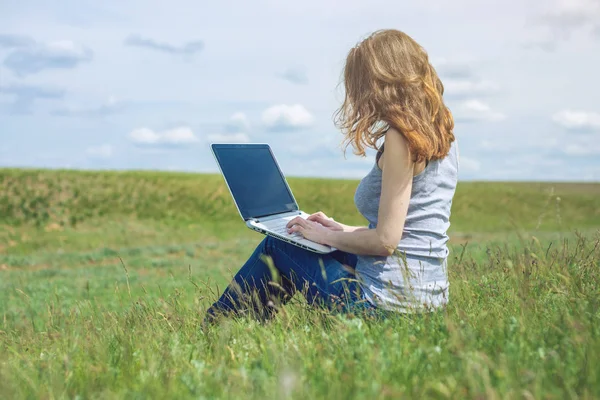 Donna seduta su un prato verde sullo sfondo del cielo con nuvole e lavorare o studiare con il computer portatile senza fili — Foto Stock