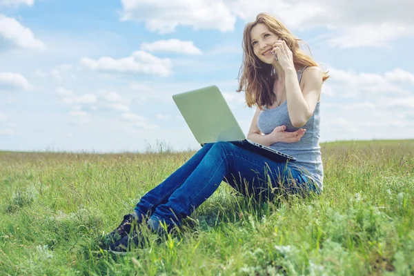 Kvinna sitter på en grön äng på bakgrunden av himlen med moln och arbetar eller studerar med bärbar dator trådlöst — Stockfoto