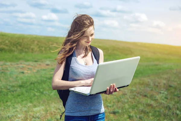 Donna in piedi su un campo verde sullo sfondo del cielo con nuvole e lavorare o studiare con il computer portatile wireless — Foto Stock