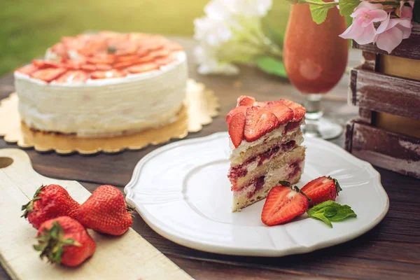 Delicious homemade summer cake with strawberries and butter cream on a wooden porch and a piece on the plate