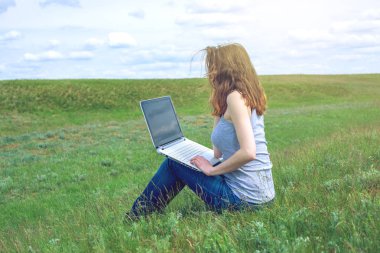 Yeşil çayır bulutlu gökyüzü arka plan üzerinde oturan ve çalışma veya kablosuz laptop ile eğitim kadın