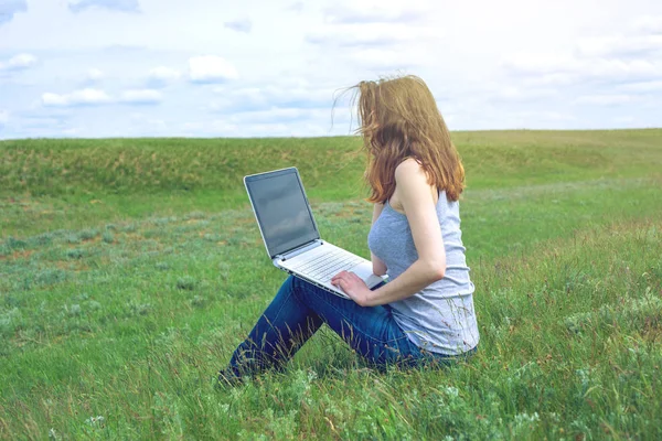 Femme assise sur une prairie verte sur le fond du ciel avec des nuages et travailler ou étudier avec un ordinateur portable sans fil — Photo