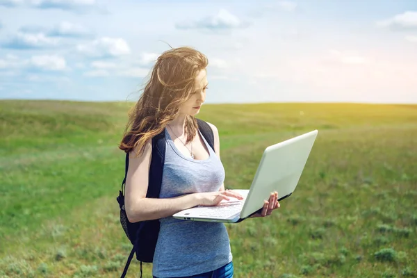 Donna in piedi su un campo verde sullo sfondo del cielo con nuvole e lavorare o studiare con il computer portatile wireless — Foto Stock