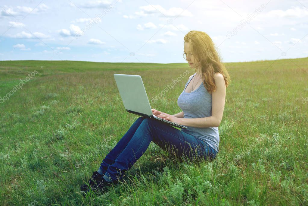Woman sitting on a green meadow on the background of sky with clouds and working or studying with laptop wireless