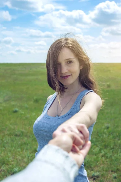 Follow me, Attractive brunette girl holding the hand of the leads in a clean green field, steppe with clouds