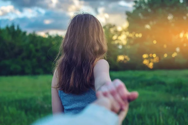 Follow me, Attractive brunette girl holding the hand of the leads in a clean green field with trees at sunset