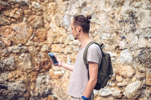 Hombre viajero con mochila mirando su teléfono inteligente en el fondo de una pared de piedra — Foto de Stock