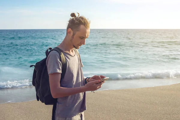 Hombre viajero con mochila mirando a su teléfono inteligente en el fondo del mar y las olas del océano . — Foto de Stock
