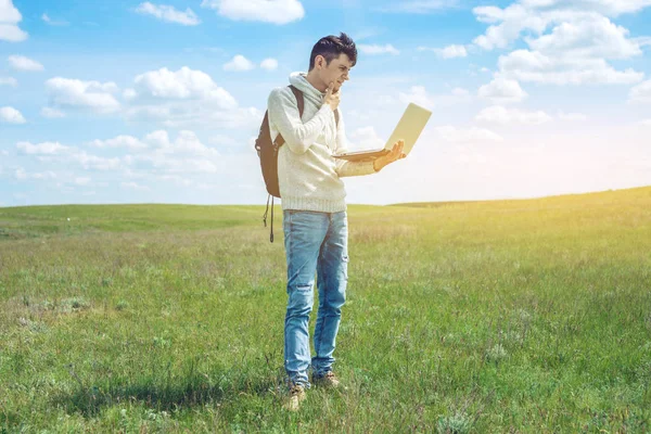 Jeune homme assis sur une prairie verte avec ordinateur portable sans fil sur le fond du ciel nuageux bleu — Photo