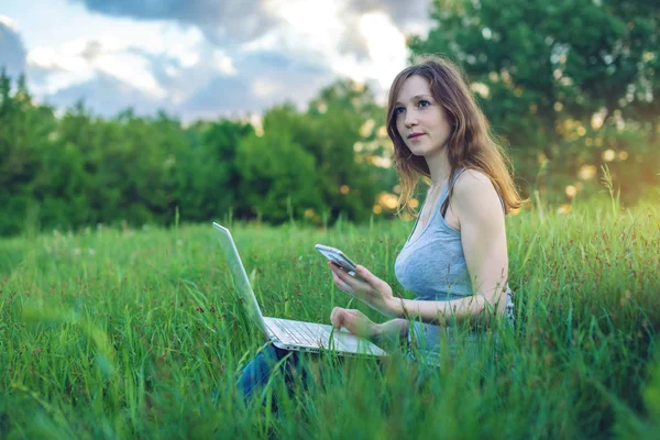 夕日の背景に緑の牧草地に座っている女性。無線と携帯電話を見てのラップトップに取り組んで. — ストック写真