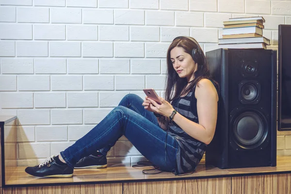 Kobieta z słuchawki słuchanie audiobooka na smartfonie siedzi na tle ściany — Zdjęcie stockowe