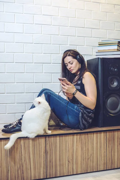 Woman słuchawki słuchanie audiobooka na telefon siedzi z kotem na tle ściany — Zdjęcie stockowe