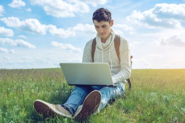 Mavi bulutlu gökyüzü arka plan üzerinde kablosuz dizüstü bilgisayar ile bir yeşil çayır üzerinde oturan genç adam