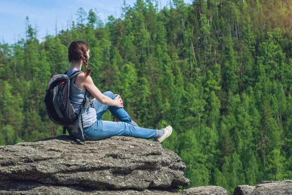 Girl with backpack sitting on a rock on top of a mountain on the background of green forest — Stock Photo, Image