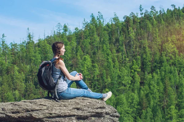 Girl with backpack sitting on a rock on top of a mountain on the background of green forest — Stock Photo, Image