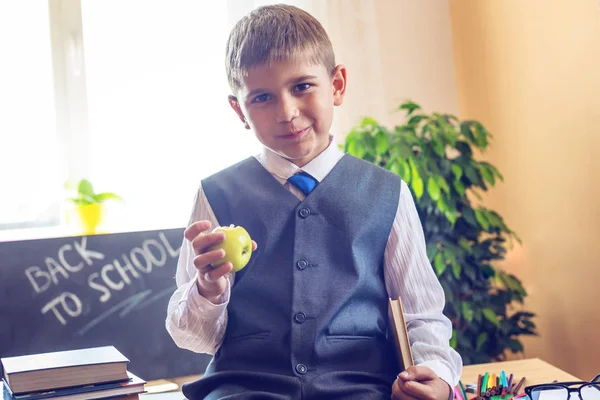 Vissza az iskolába. Aranyos gyerek ül a tanteremben a íróasztal. Fiú eszik almát, iskolai szünetet — Stock Fotó