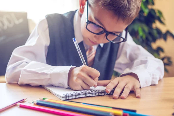 De volta à escola. Criança bonita sentada na mesa da sala de aula. Menino está aprendendo lições escreve uma caneta em um caderno — Fotografia de Stock