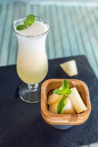 Batido blanco fresco en un vaso con trozos de melón y menta en rodajas sobre un fondo de madera azul. Bebidas frescas de verano — Foto de Stock