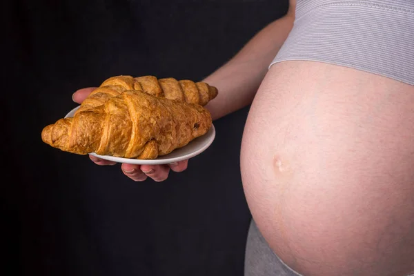 Egy terhes nő hasa gazdaság egy lemezt a croissant. Koncepció súlykontroll és az egészségtelen táplálkozás, a terhesség alatt — Stock Fotó