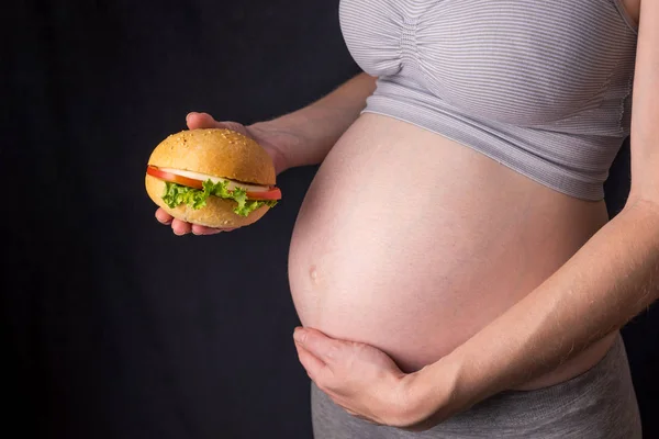 Uma mulher grávida com barriga a segurar um hambúrguer. Conceito de fast food e alimentação não saudável durante a gravidez — Fotografia de Stock