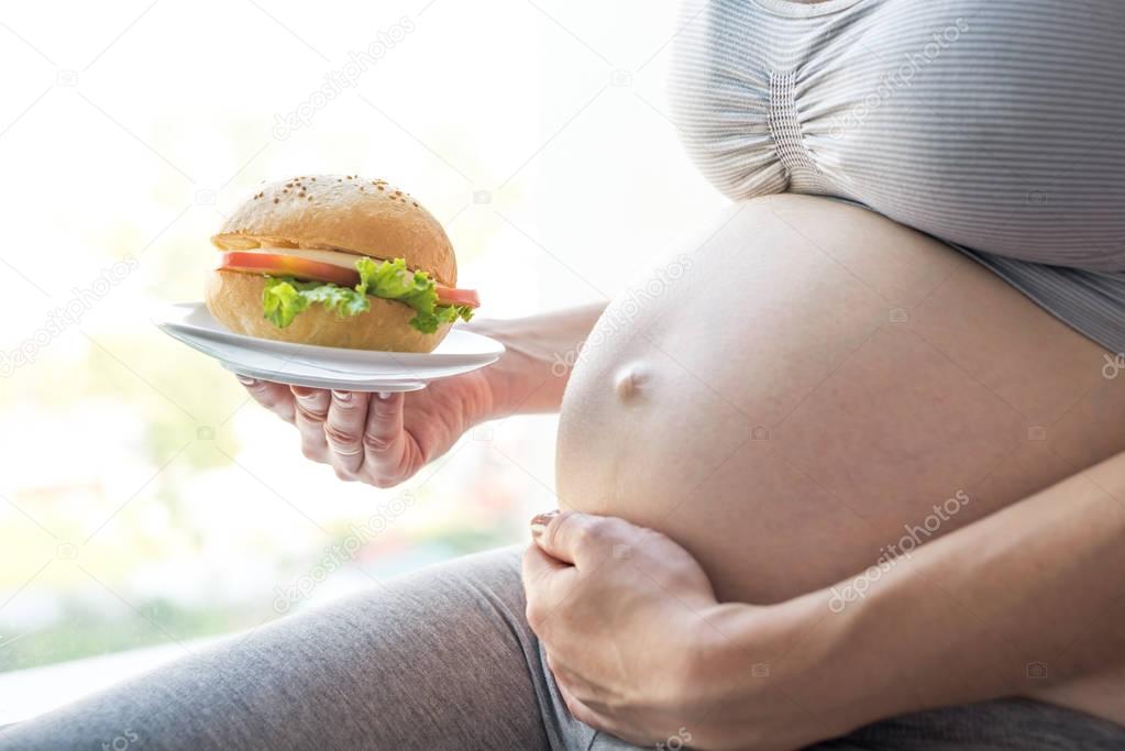 A pregnant woman with belly by the window holding a burger. Concept of fast food and unhealthy eating during pregnancy