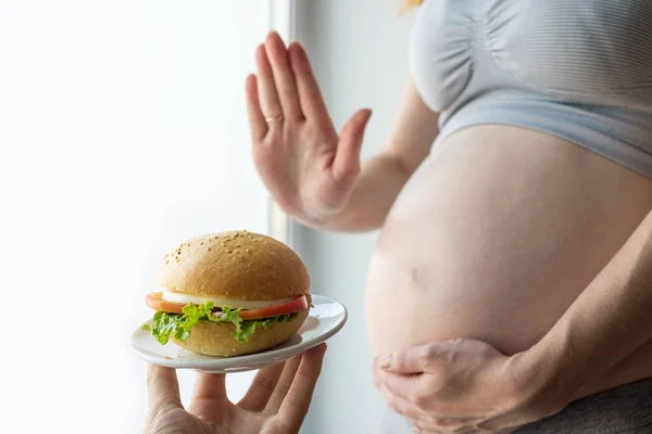 Ciężarna kobieta z brzuchem przy oknie odmawia burgera. Kontrola koncepcji podczas ciąży. Fast food — Zdjęcie stockowe