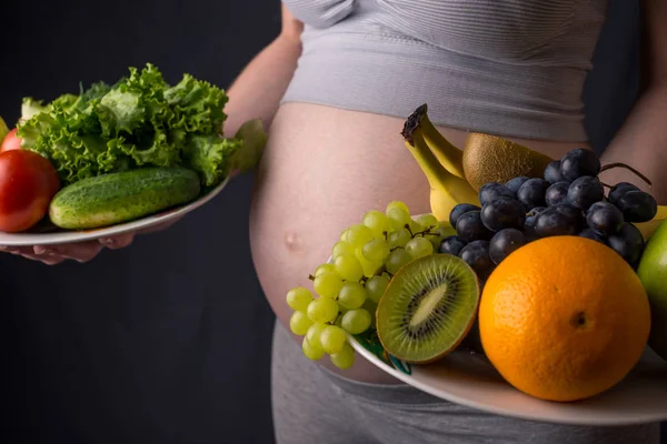 Terhes nő hasa egy tányér, a gyümölcsök és zöldségek tartja kezében. Koncepció egészséges táplálkozás a terhesség alatt — Stock Fotó