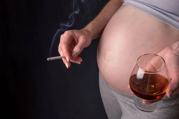 Seorang wanita hamil dengan perut memegang segelas wiski dan rokok. Konsep alkohol dan kebiasaan buruk selama kehamilan Stok Foto Bebas Royalti