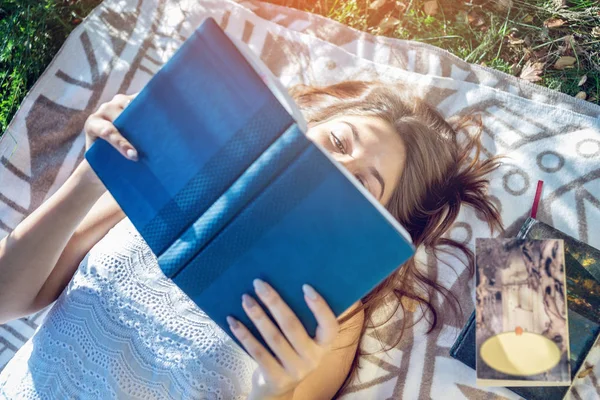 Mujer leyendo un interesante libro que yace en el parque en un césped verde en un día soleado de verano. Concepto de educación de los estudiantes —  Fotos de Stock
